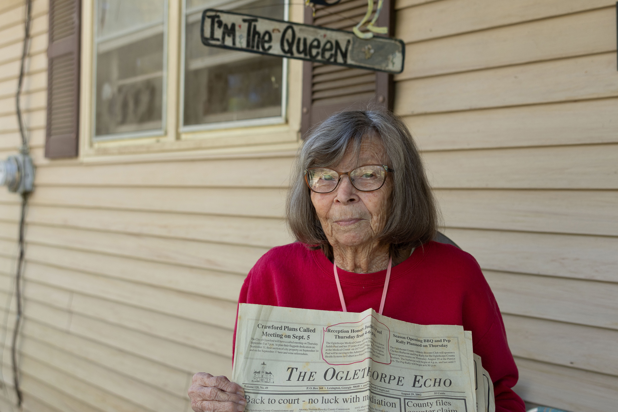 Judith Paul, 85, poses for a portrait at her home located in the community of Sandy Cross in Oglethorpe County, Georgia on March 27, 2025. She holds a 2002 print copy of The Oglethorpe Echo that recognizes her 25 years of service as the first secretary of Oglethorpe Primary Health Care Center, the first ever healthcare facility to open in Crawford, Georgia in 1979. Above her is a sign that reads "I'm The Queen," made for her by her two sons, Jay and Lee Paul, in honor of her nickname at the Medical Center, 