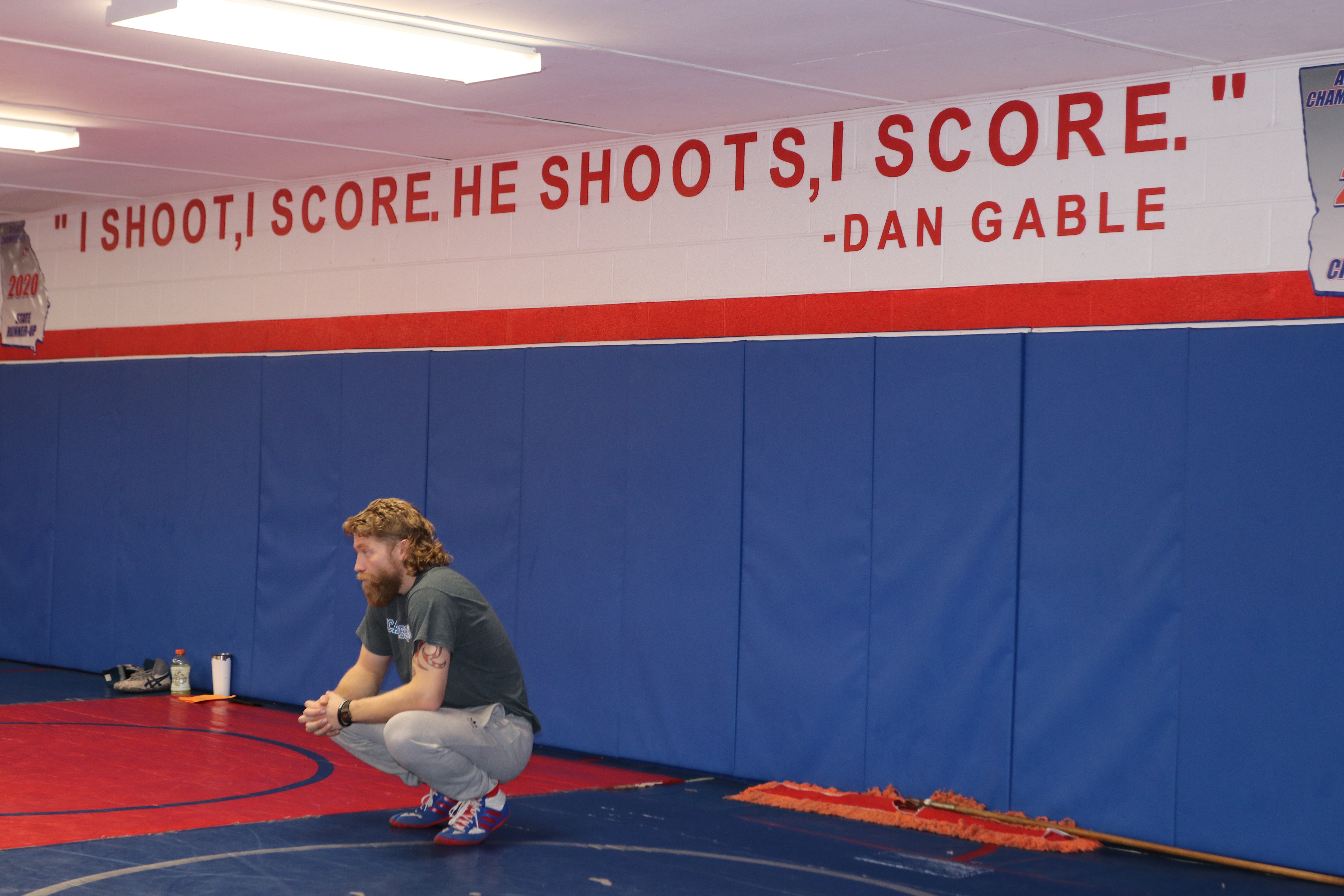 Oglethorpe County Middle School head wrestling coach Cory Collins squats down during a practice at the Oglethorpe County High School gymnasium. Above him, a famous quote from wrestling icon Dan Gable defines the aggressive, competitive mindset of the program. (Photo/Grant Turnage) 