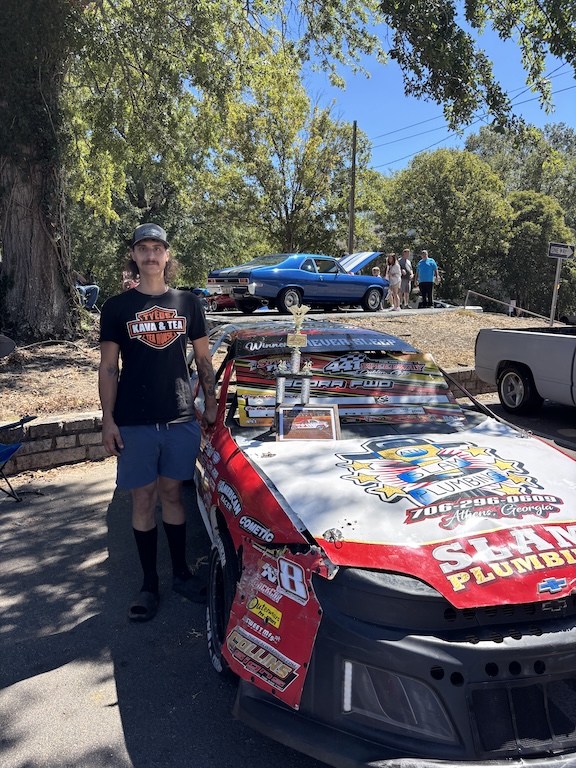 Luke Lackore stands next to his car at the Firefly Cruise In on Sunday at the courthouse in Lexington. Lackore built the car, with help from his family, and covered it in advertisements for local sponsors that he works with at his plumbing job. (Maura Potvin/The Oglethorpe Echo)