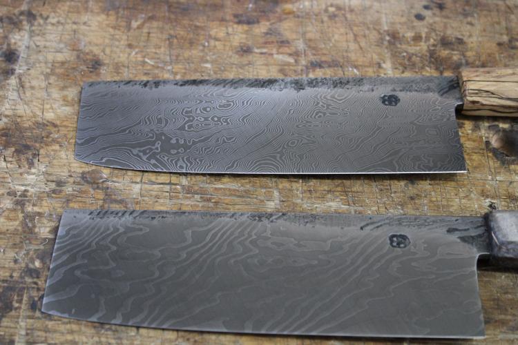 A ground coffee patina finish and embossed logo are visible on two finished cleaver knives on Feb. 13, 2023. The slightly acidic coffee combined with ferric chloride bring out a distinct finish that both protects the knife and pleases the eye. (Photo/Sarah Donehoo)