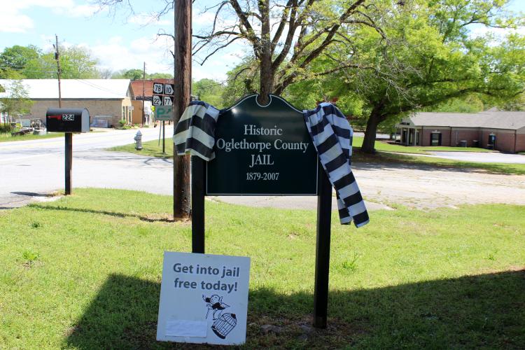 The Oglethorpe County Historic Jail hosts its first art exhibit featuring Tom Thon’s artwork on Saturday, April 12. Fellow artist and jail custodian John Fortuna attended to support Thon, posting a sign for passersby that reads, “Get into jail free today.” (Photo/Emily Slepsky)