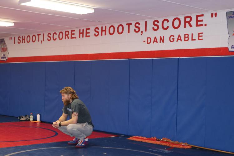 Oglethorpe County Middle School head wrestling coach Cory Collins squats down during a practice at the Oglethorpe County High School gymnasium. Above him, a famous quote from wrestling icon Dan Gable defines the aggressive, competitive mindset of the program. (Photo/Grant Turnage)