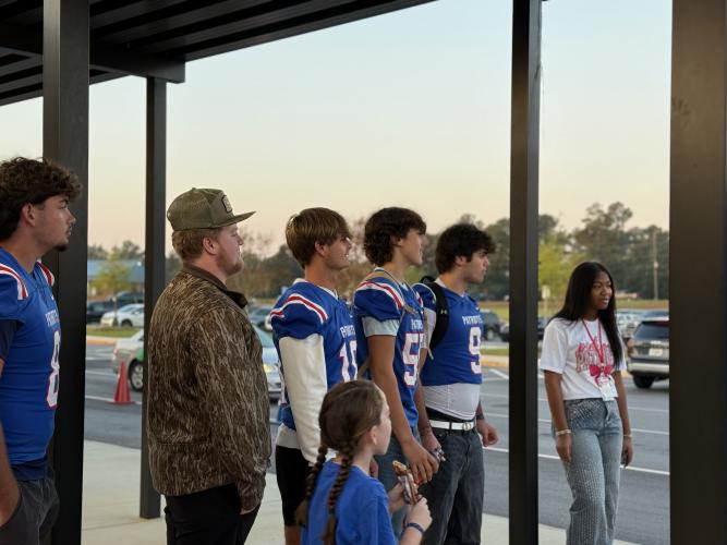Football players Brett Liedberg, JD Armour, Justin Wilson, Lance Ellefson and Coleton Sexton, along with cheerleader Nevaeh Calhoun, stand in line, awaiting student drop-offs. “I think they’re like expecting it every Friday, or like almost every Friday, and they, like, get excited,” said Calhoun about the impact the weekly greeting has on the kids. (Photo/Walker Durham)