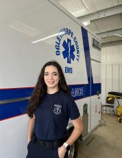Kate Spearing, a 2023 Oglethorpe County High School graduate, stands with a county ambulance in her emergency medical services uniform. Spearing has logged more than 200 clinical hours as an EMT and will compete at HOSA’s International Leadership Conference in Dallas starting June 21. (McCain Bracewell/The Oglethorpe Echo)