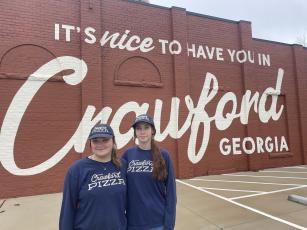 Crawford Pizza employees Elizabeth Russell (left) and  Caitlin Hartrum stand in front of the restaurant’s mural  earlier this month. Russell, who lives in Crawford, and  Hartrum, who lives in Lexington, said it helps welcome  people to the county. (Margaux Binder/The Oglethorpe Echo)