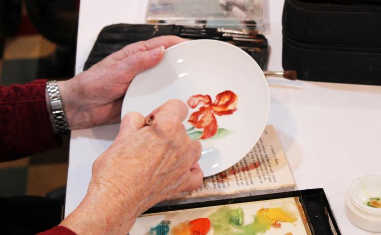 Jane Bowen paints a flower on a plate in her studio in Crawford.  She also paints lamps, furnishings, tiles and dishes. (Shannon Moran/The Oglethorpe Echo)